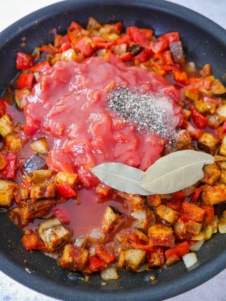 A frying pan filled with sauteed and spiced vegetables with added canned plum tomatoes, bay leaves, salt and black pepper.