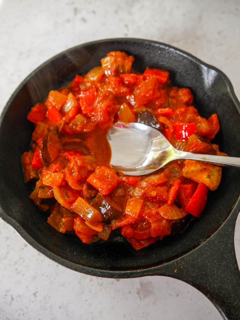 A cast iron skillet filled with sauteed aubergine, peppers, onion and garlic in a spicy tomato sauce.