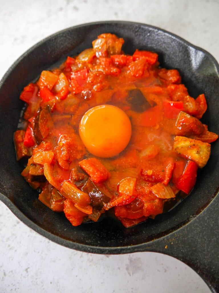 A cast iron skillet filled with sauteed aubergine, peppers, onion and garlic in a spicy tomato sauce with an egg cracked on top.
