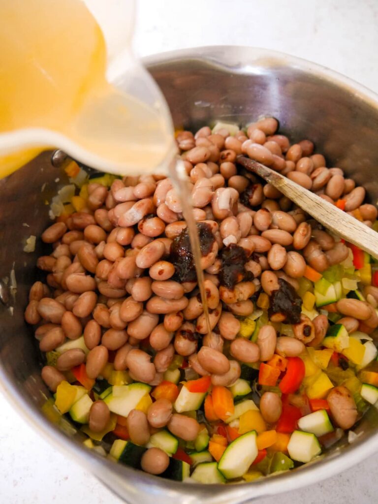 A large saucepan with sauteed vegetables with added borlotti beans, chipotle chilli paste and vegetable stock being poured over.