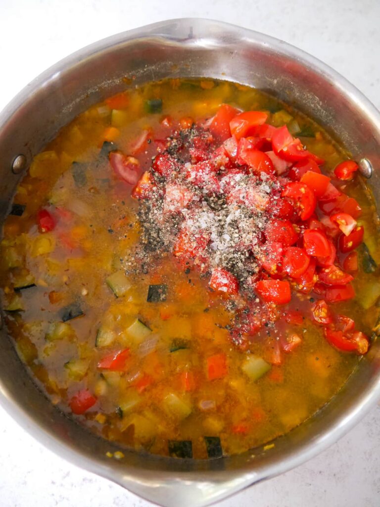 A large saucepan of soup with fresh tomatoes, salt and black pepper being added.