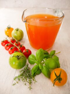 A glass jug filled with fresh tomato stock, with fresh tomatoes, basil and thyme leaves set alongside.