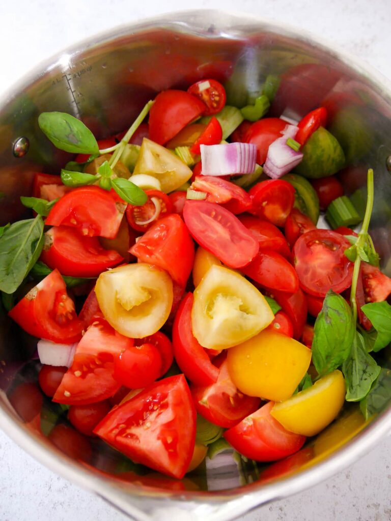 A large saucepan filled with chopped tomatoes, red onion, celery, sprigs of thyme and basil.