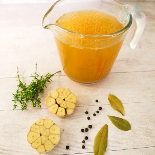 A jug of homemade chicken stock with fresh thyme, garlic, black peppercorns and dried bay leaves set alongside.