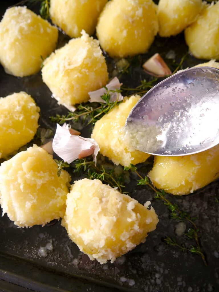 Par boiled potatoes added to a roasting dish with garlic and thyme, with oil being spooned over the potatoes.