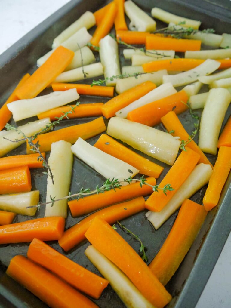 A roasting dish with carrot and parsnip batons covered in oil and honey with added sprigs of thyme.