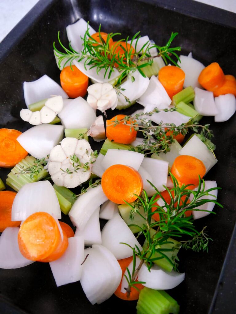 A roasting dish filled with raw vegetables and fresh herbs, including onion, carrot, celery, garlic, thyme and rosemary.