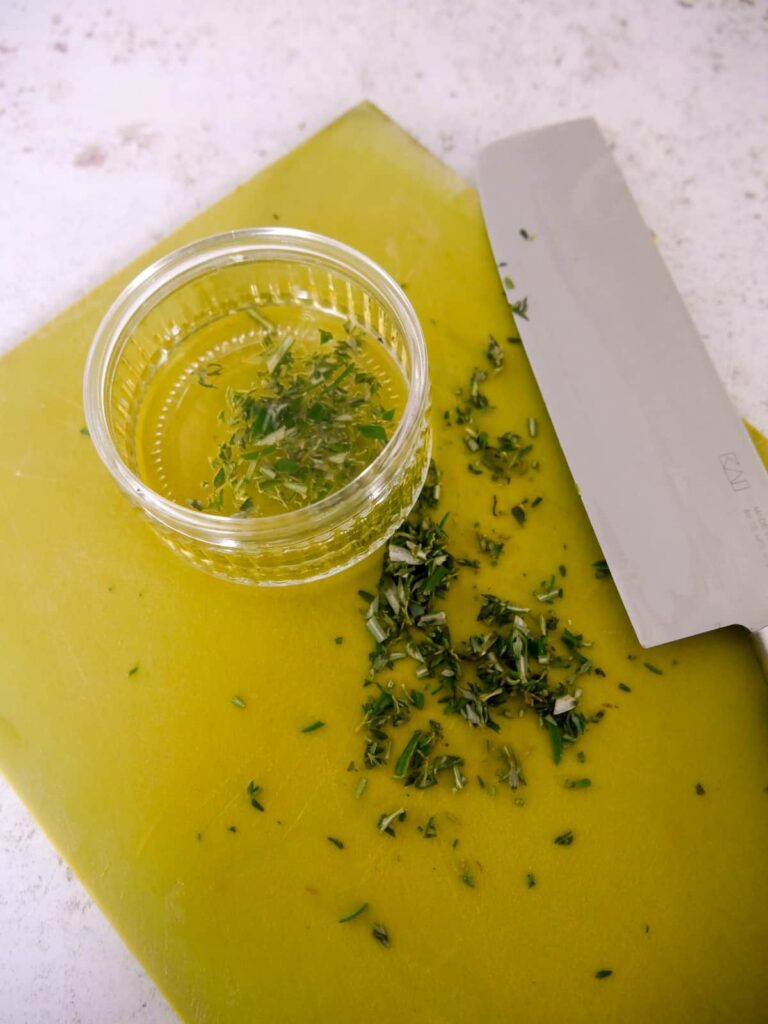 A chopping board with chopped rosemary and thyme with a bowl of herb infused olive oil set alongside.