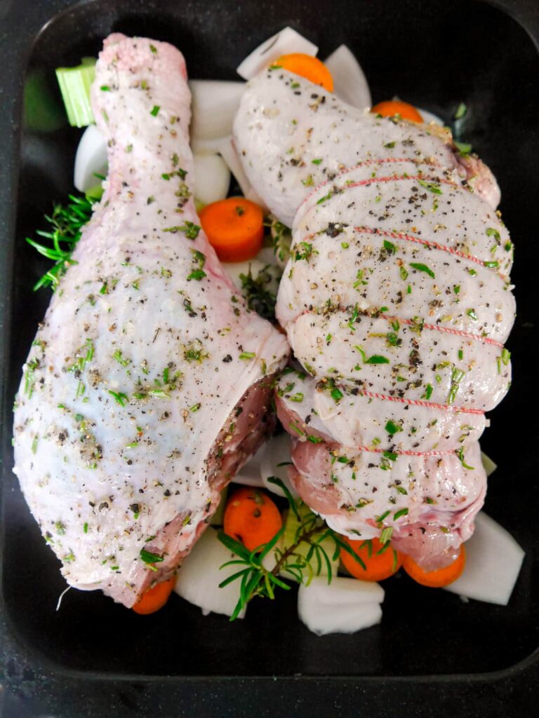 A roasting dish with seasoned turkey drumstick and thigh set on top of vegetables, ready for the oven.