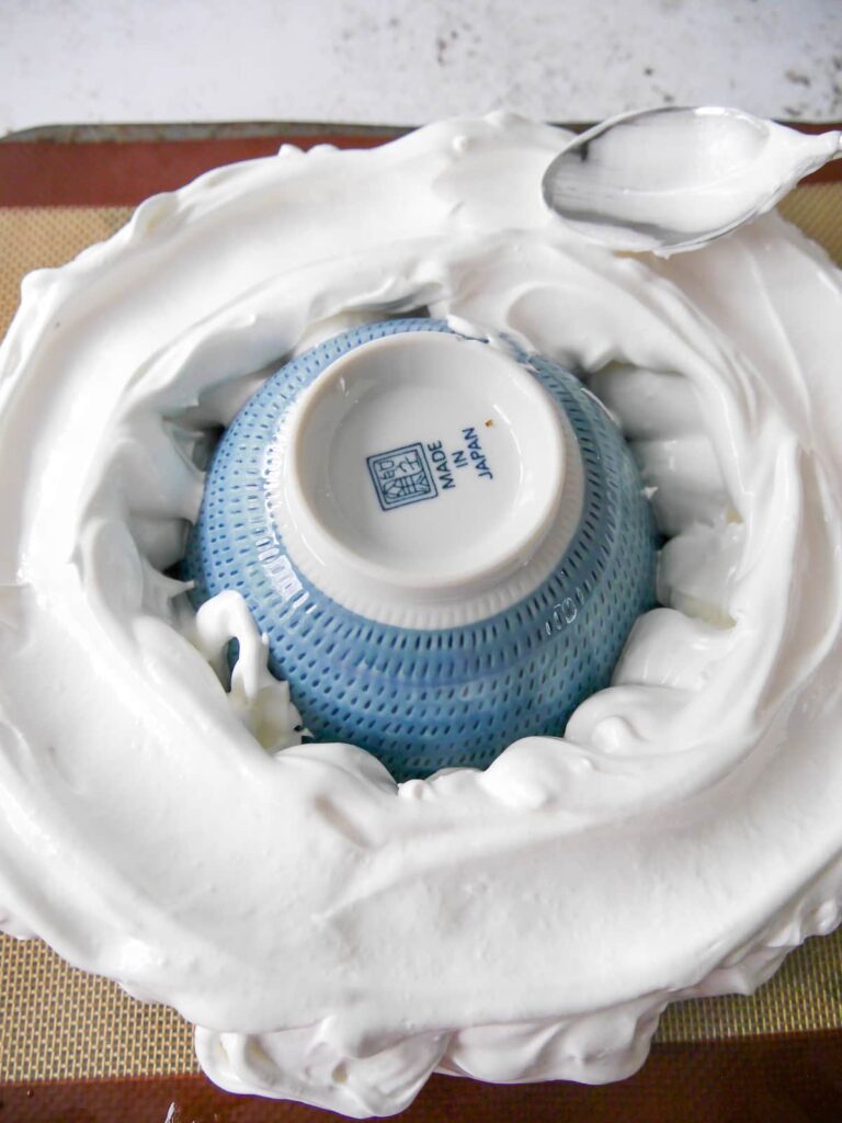 A baking sheet topped with a ring of meringue with a bowl in the centre and a spoon making a well across the top of the meringue.