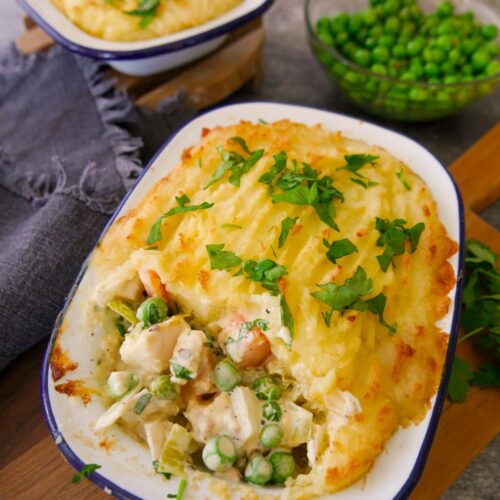 Two dishes of creamy fish pie topped with mashed potatoes and garnished with chopped parsley, with a bowl of garden peas set alongside.