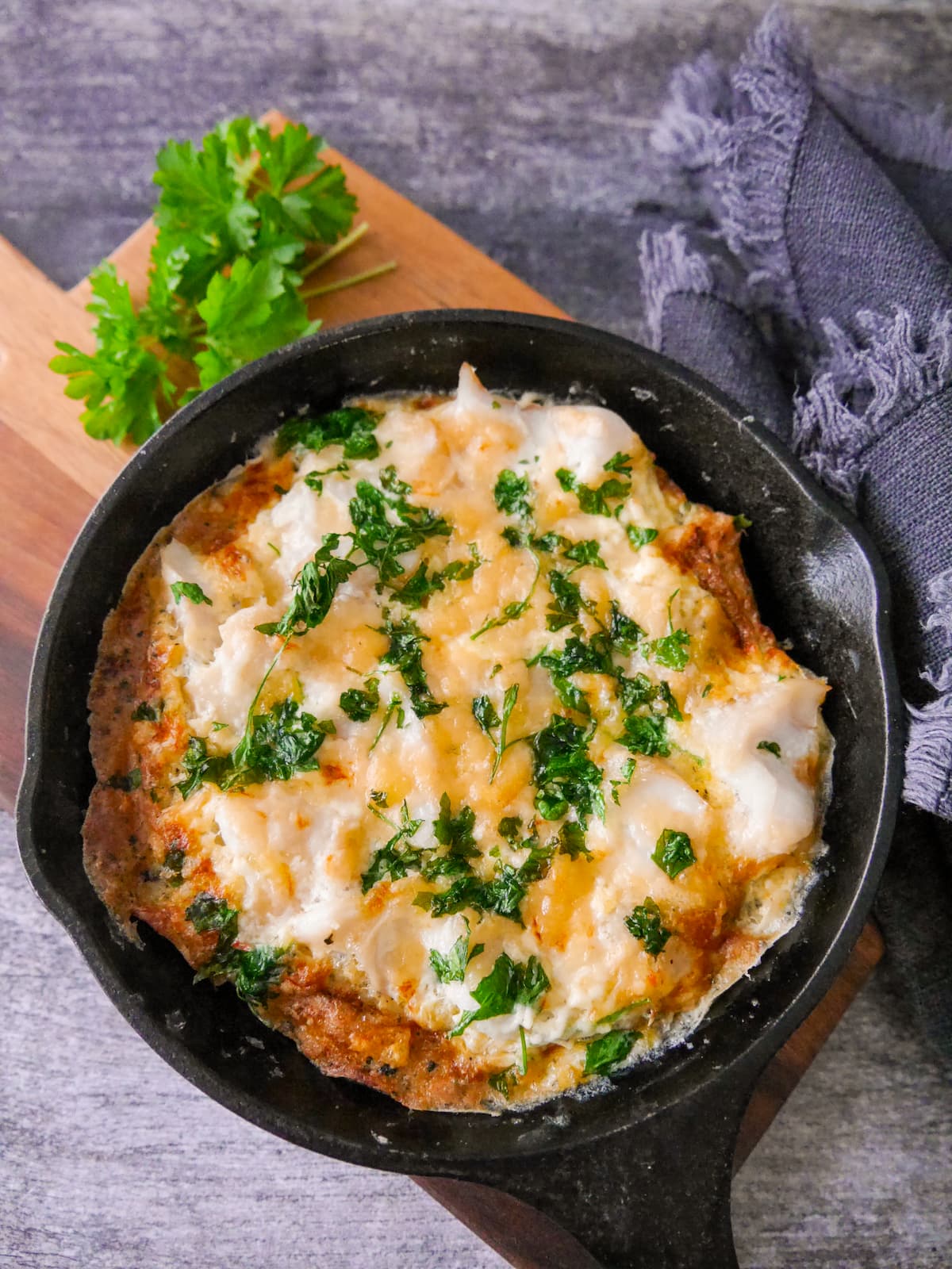 A cast iron skillet filled with omelette Arnold Bennett and topped with chopped parsley, set on a wooden board.