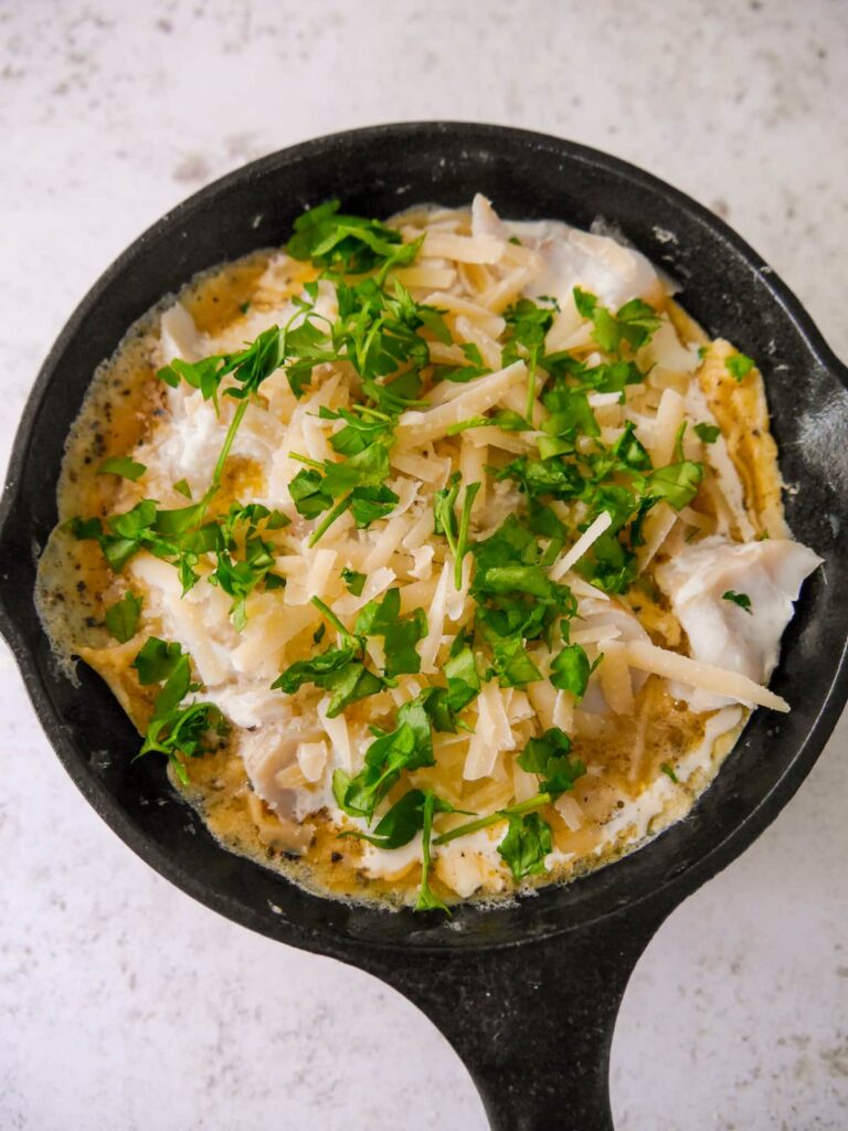 A cast iron skillet with a cooked omelette base topped with pieces of smoked haddock, some grated parmesan cheese and chopped parsley.