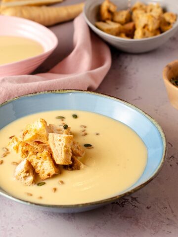 A bowl of creamy parsnip soup garnished with croutons and toasted pumpkin and sunflower seeds.