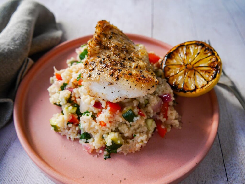 Pan fried hake fillet sitting on top of a plate of lemon and vegetable couscous. served with half a burnt lemon on the side.