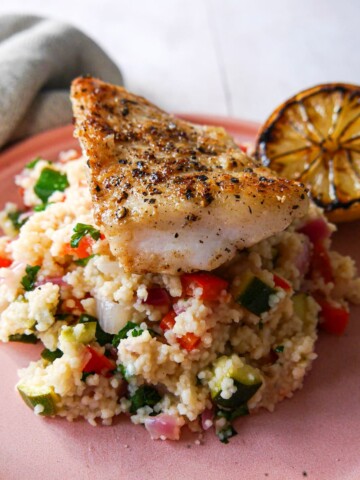 Pan fried hake fillet sitting on top of a plate of lemon and vegetable couscous. served with half a burnt lemon on the side.