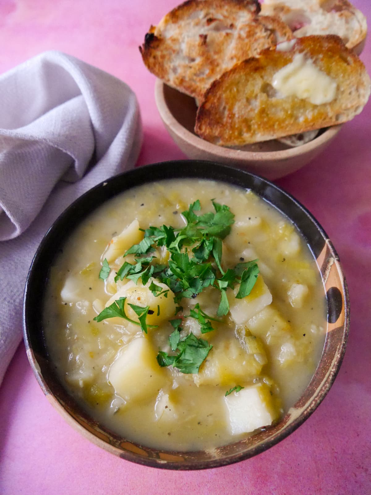 A bowl of rustic leek and potato soup topped with a garnish of chopped parsley, with a side of buttered toast.