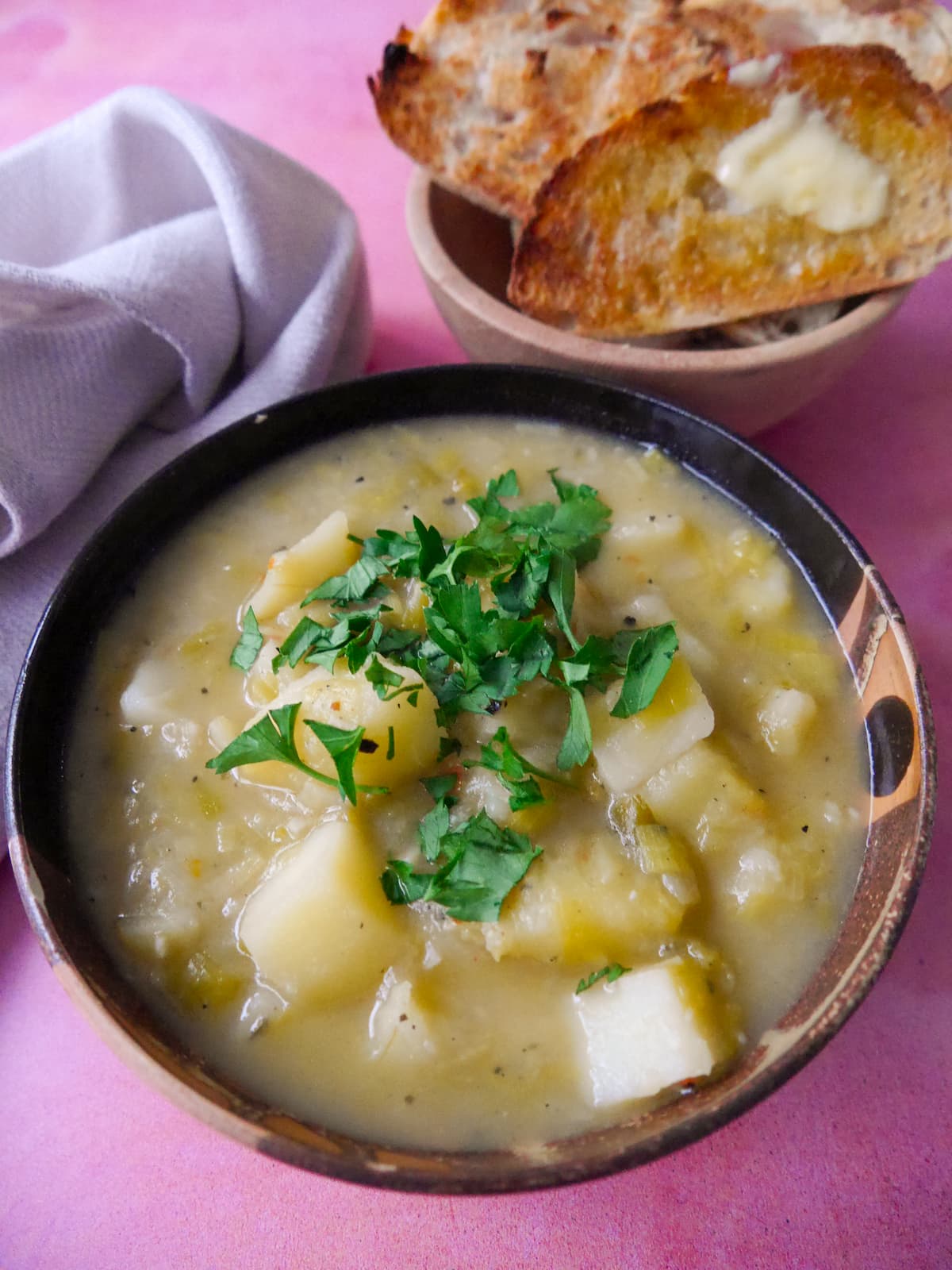 A bowl of rustic leek and potato soup topped with a garnish of chopped parsley, with a side of buttered toast.