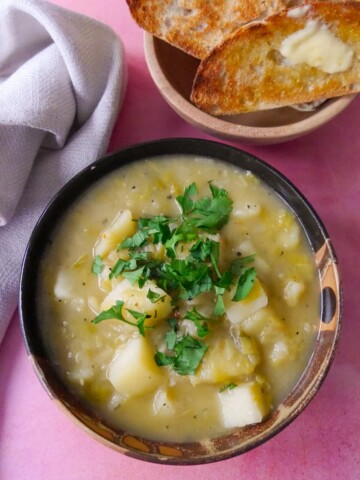 A bowl of rustic leek and potato soup topped with a garnish of chopped parsley, with a side of buttered toast.