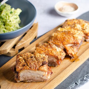 Slices of roast pork belly with crispy crackling set on a wooden board, with a side of creamed brussels sprouts.