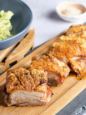 Slices of roast pork belly with crispy crackling set on a wooden board, with a side of creamed brussels sprouts.