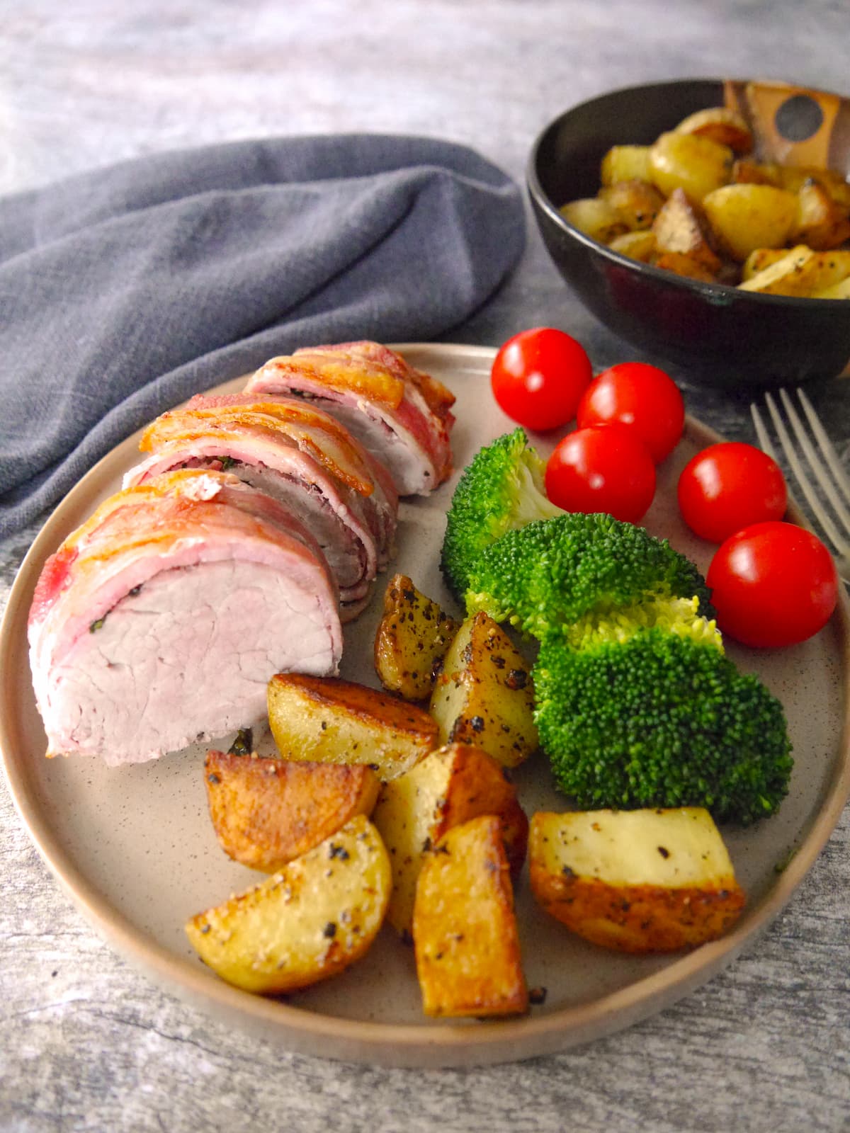 A plate with slices of bacon wrapped pork fillet set on top and served with sauteed potatoes, steamed broccoli and fresh tomatoes.