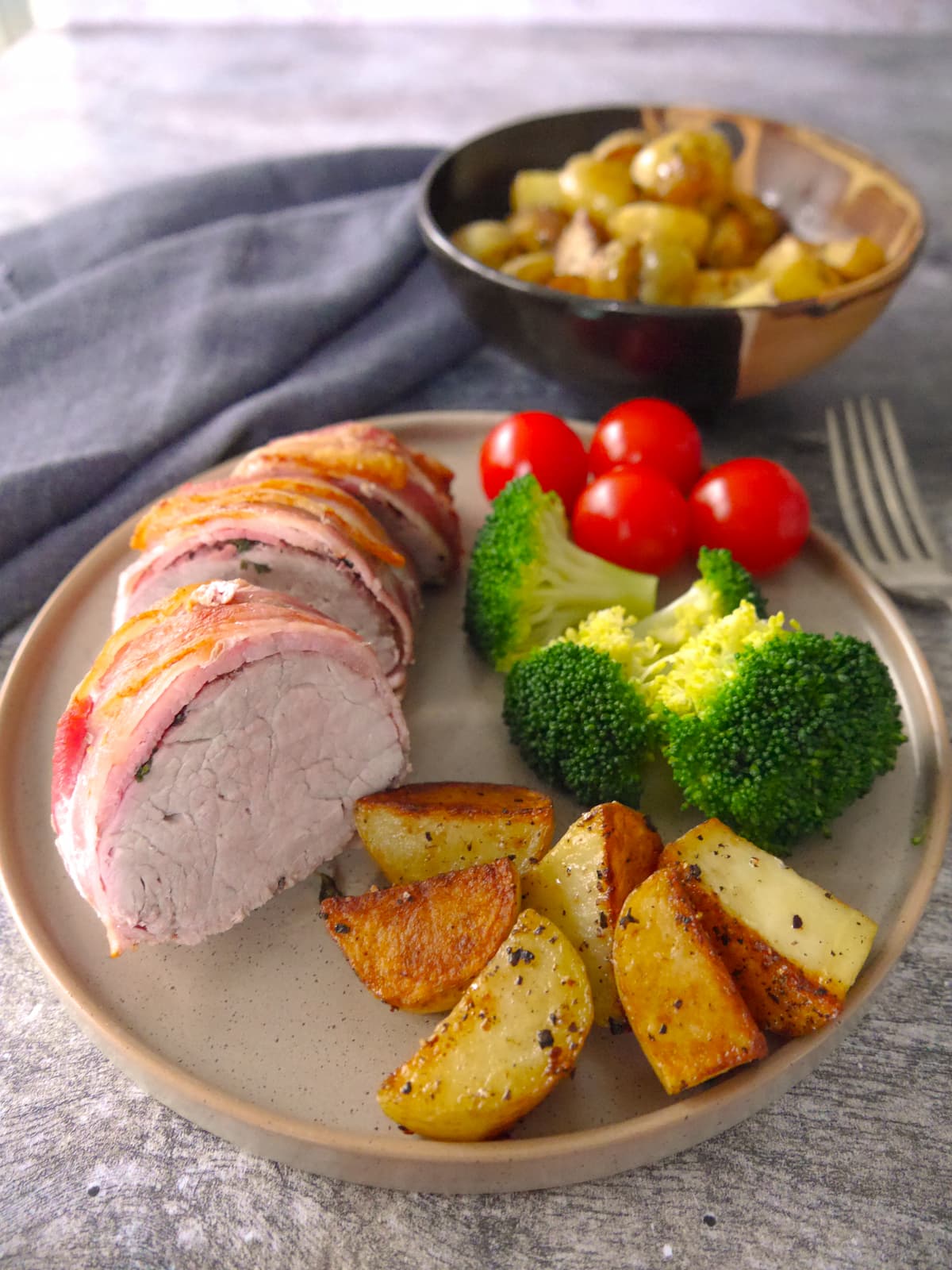 A plate with slices of bacon wrapped pork fillet set on top and served with sauteed potatoes, steamed broccoli and fresh tomatoes.