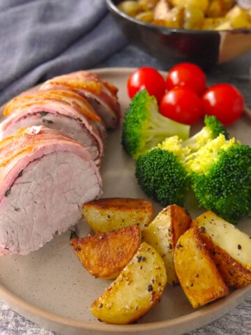 A plate with slices of bacon wrapped pork fillet set on top and served with sauteed potatoes, steamed broccoli and fresh tomatoes.