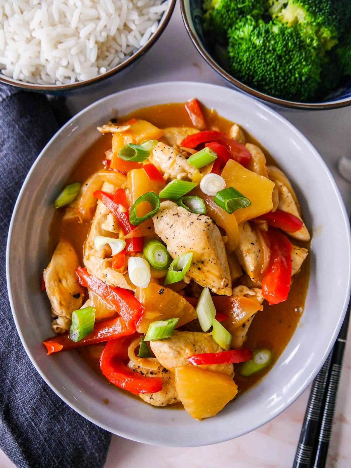 A bowl of sweet and sour chicken with stir-fried vegetables, with sides of steamed rice and broccoli.