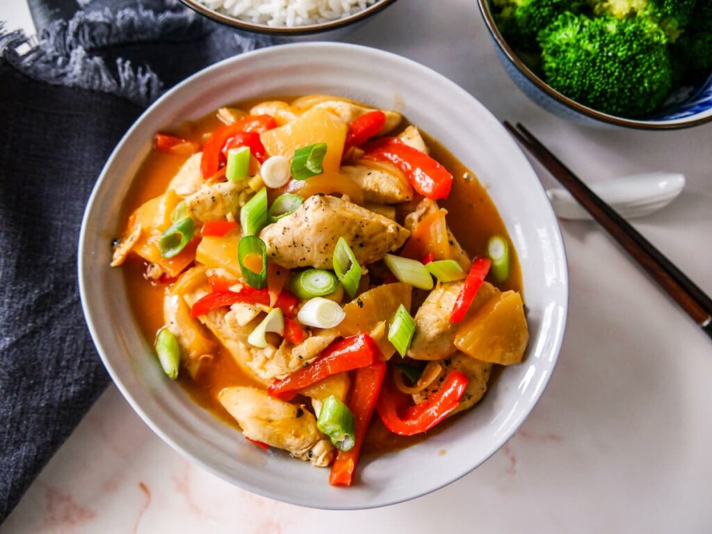 A bowl of sweet and sour chicken with stir-fried vegetables, with sides of steamed rice and broccoli.