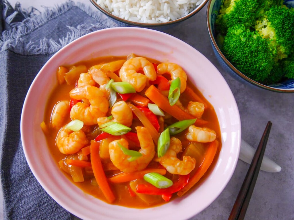 A pink bowl filled with sweet and sour prawn stir-fry, with sides of steamed rice and broccoli.