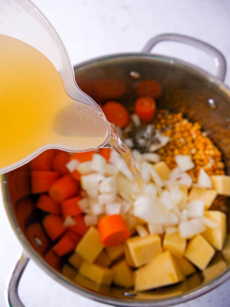 A large saucepan filled with yellow split peas, sliced carrots, chopped swede, onion and seasoning, with vegetable stock being added.