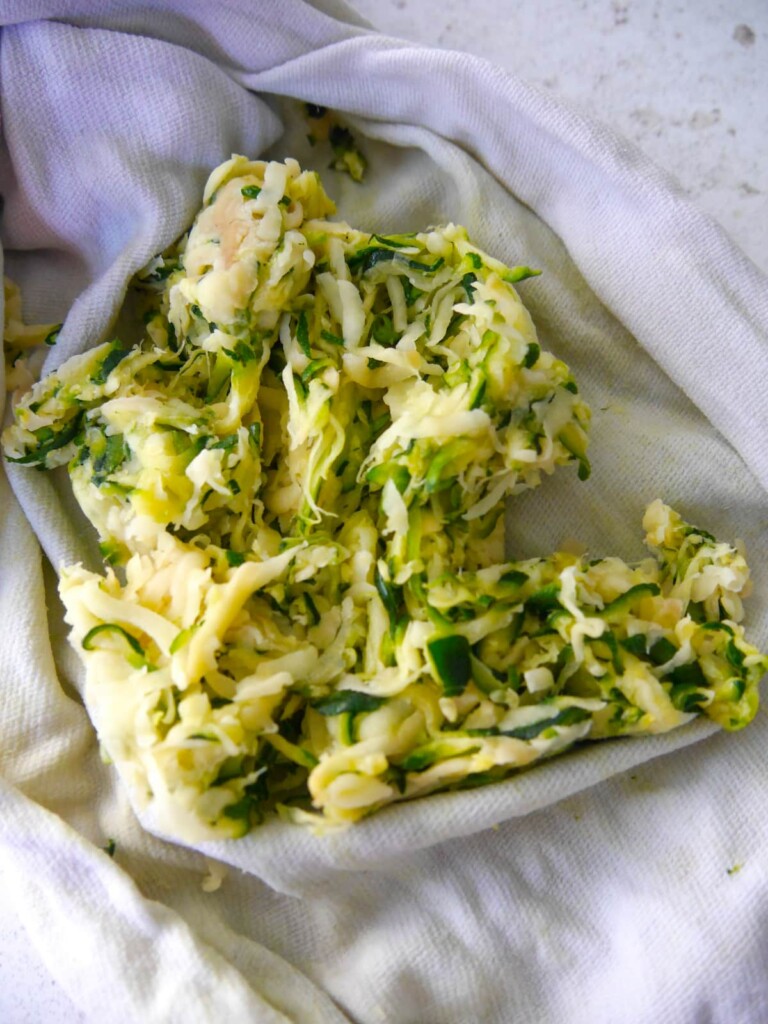 Grated courgette and potato being squeezed in a clean kitchen towel to remove excess water.