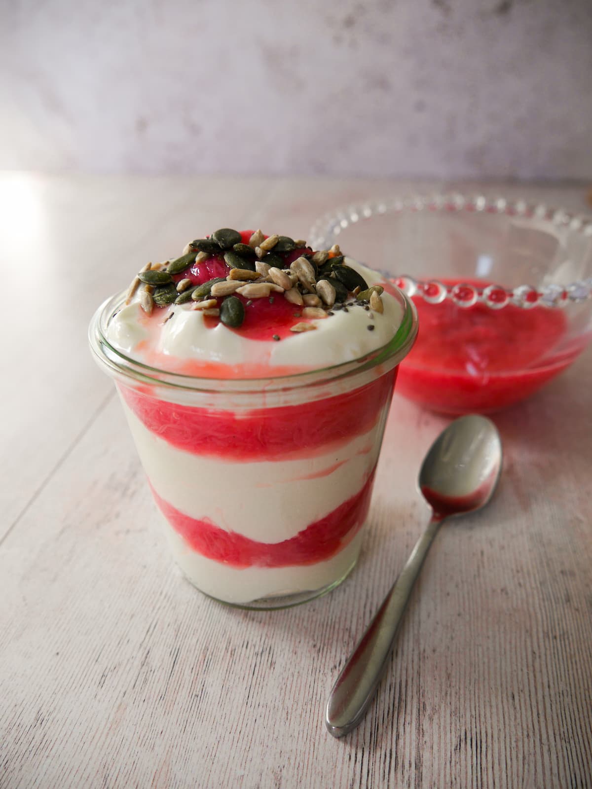 A glass filled with Greek yogurt layered with stewed rhubarb and topped with pumpkin, sunflower and chia seeds, with a bowl of healthy stewed rhubarb set alongside.