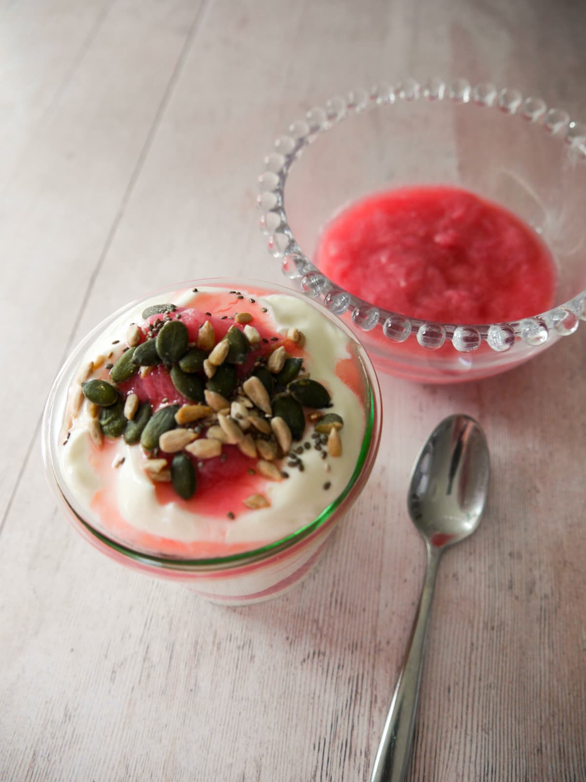 A glass filled with Greek yogurt layered with stewed rhubarb and topped with pumpkin, sunflower and chia seeds, with a bowl of healthy stewed rhubarb set alongside.