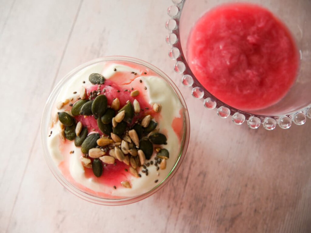 A glass filled with Greek yogurt layered with stewed rhubarb and topped with pumpkin, sunflower and chia seeds, with a bowl of healthy stewed rhubarb set alongside.