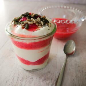 A glass filled with Greek yogurt layered with stewed rhubarb and topped with pumpkin, sunflower and chia seeds, with a bowl of healthy stewed rhubarb set alongside.