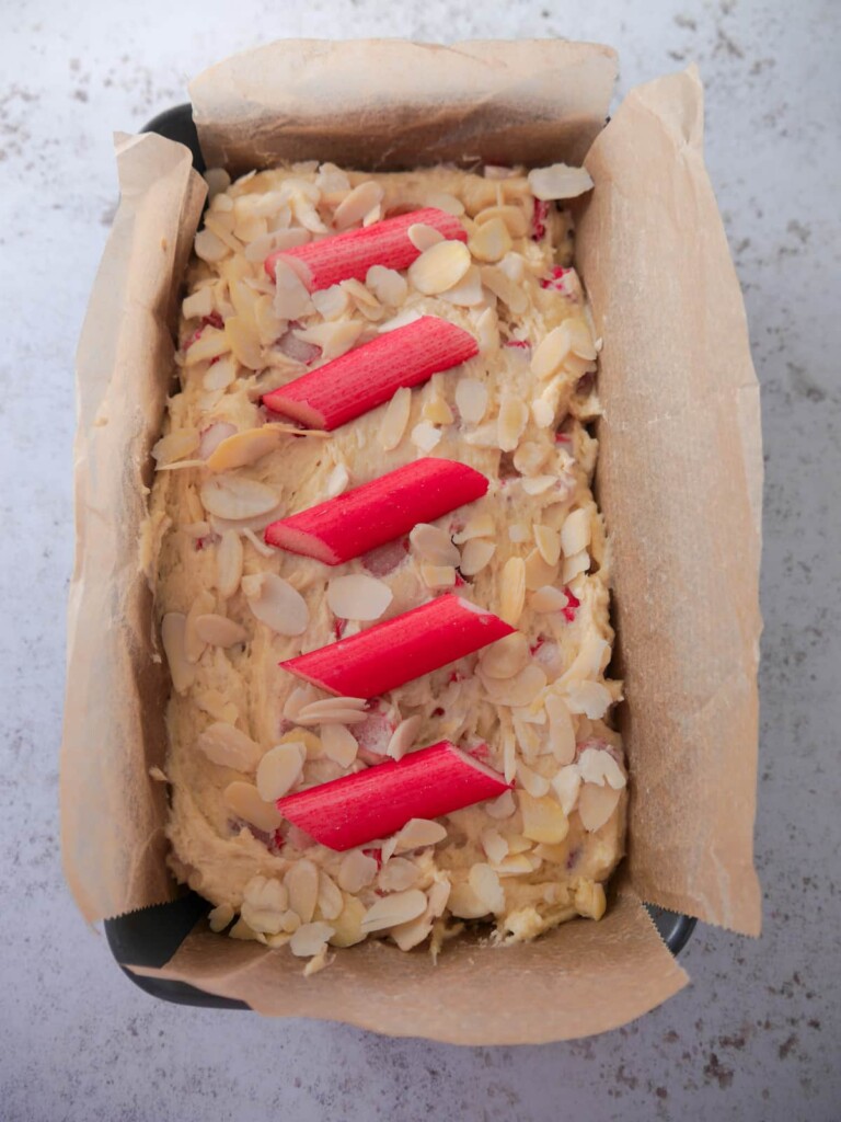Lined baking tin filled with rhubarb and almond cake batter, topped with batons of fresh rhubarb and flaked almonds.