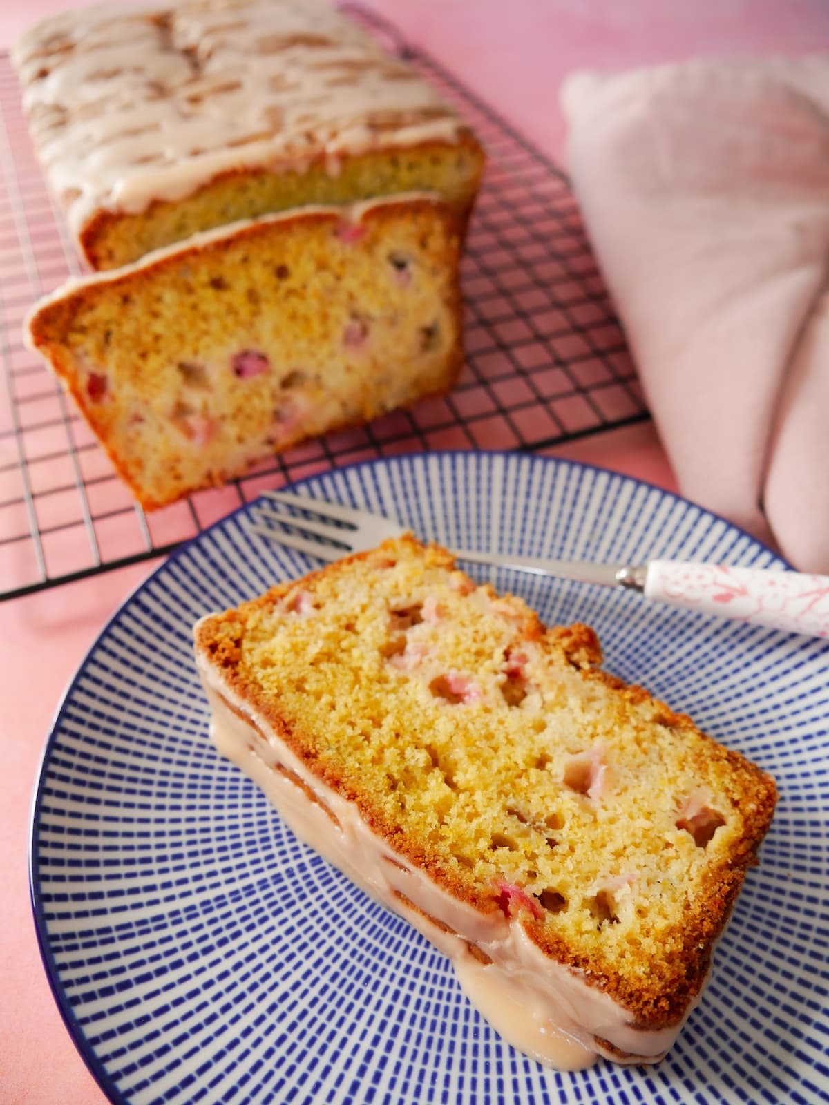 A slice of rhubarb and orange loaf cake on a plate, with the whole loaf set on a wire rack alongside.