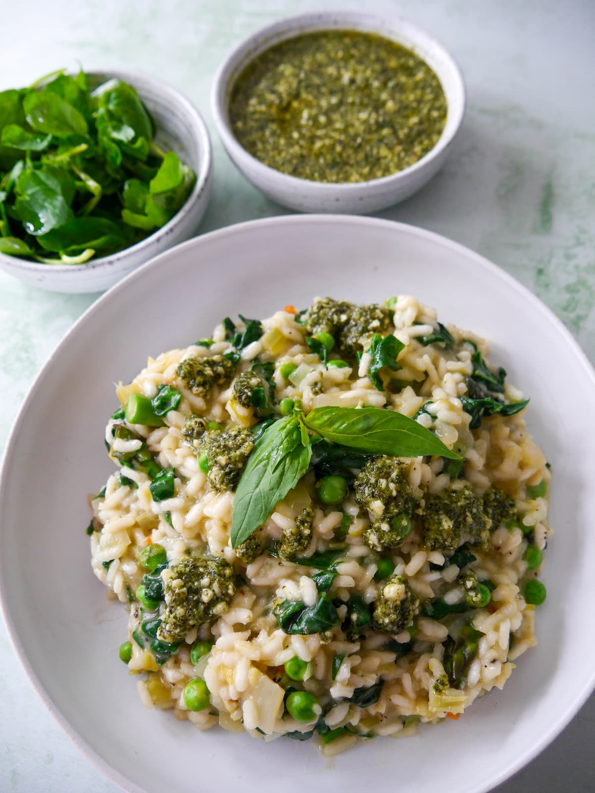 A white bowl of spring vegetable risotto, topped with a drizzle of basil pesto and garnished with fresh basil leaves, with bowls of dressed watercress salad, and pesto set alongside.