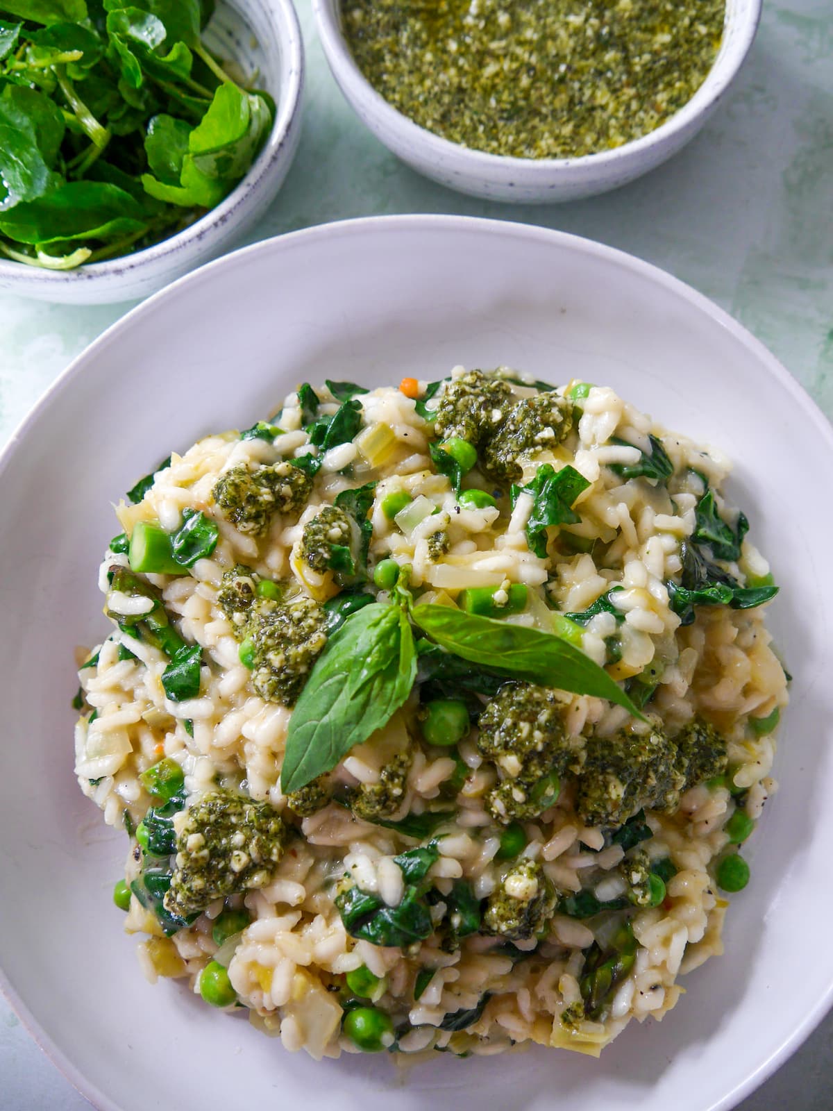 A white bowl of spring vegetable risotto, topped with a drizzle of basil pesto and garnished with fresh basil leaves, with bowls of dressed watercress salad, and pesto set alongside.