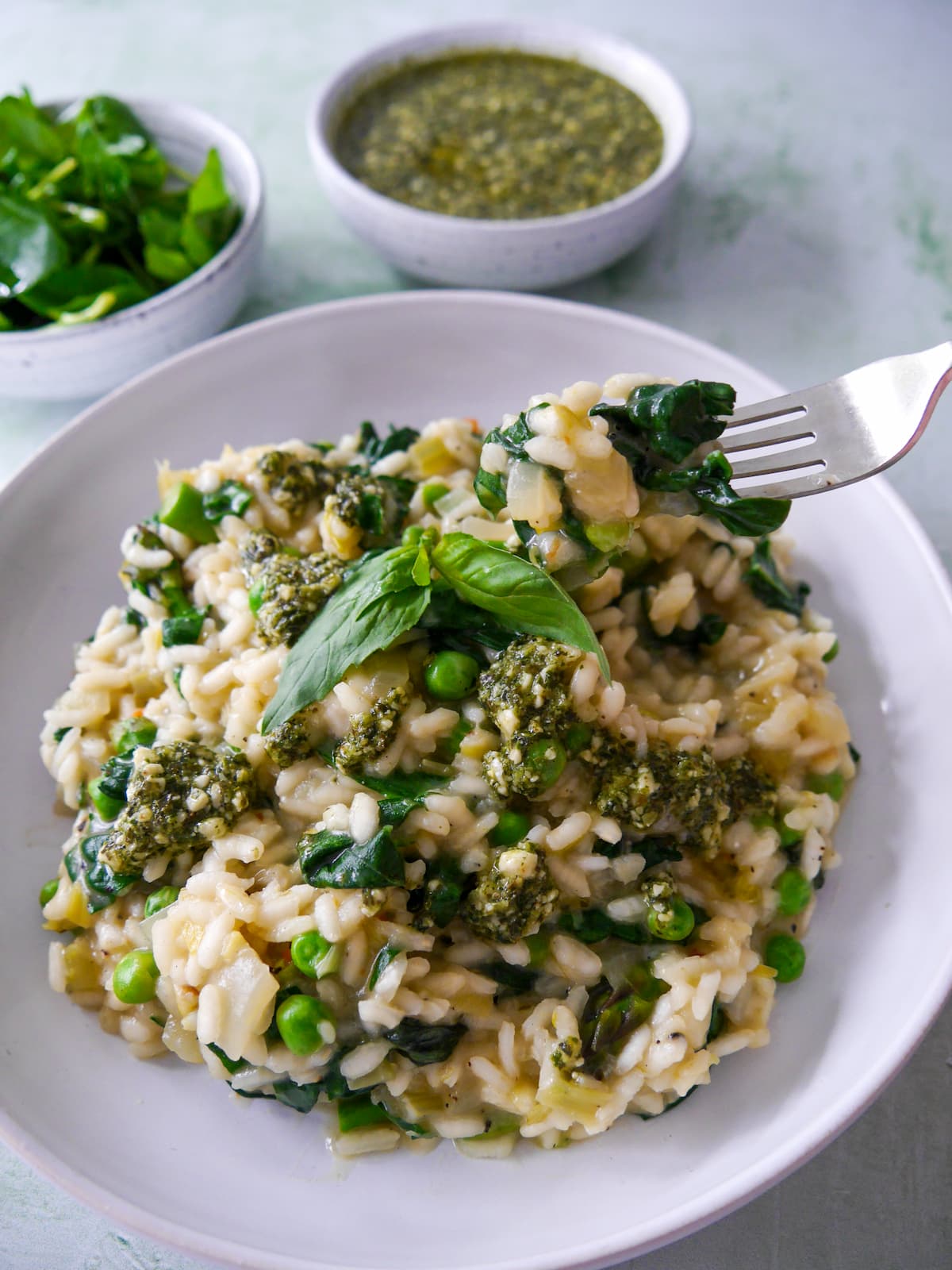 A white bowl of spring vegetable risotto, topped with a drizzle of basil pesto and garnished with fresh basil leaves, with bowls of dressed watercress salad, and pesto set alongside.