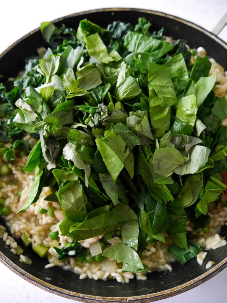 A frying pan with a base of sauteed vegetables and risotto rice, with chopped spinach, cavolo nero kale and fresh basil leaves being added.