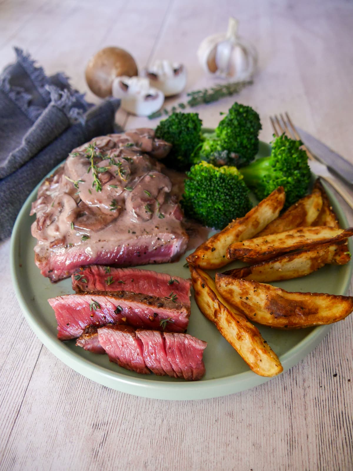 A plate of medium rare sirloin steak, topped with a red wine mushroom sauce and served with a side of potato wedges and steamed broccoli.