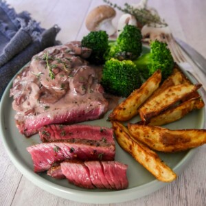 A plate of medium rare sirloin steak, topped with a red wine mushroom sauce and served with a side of potato wedges and steamed broccoli.