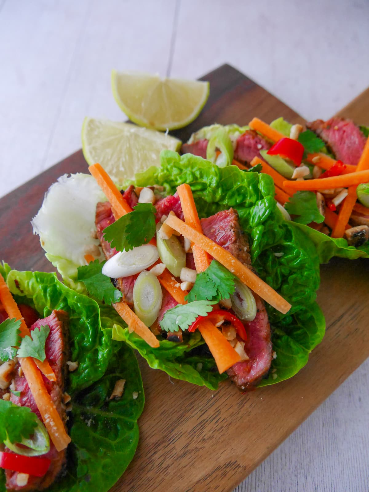 A wooden board topped with 3 lettuce leaves, laden with sliced lemongrass steak strips, crunchy carrot strips, spring onion, crushed peanuts and coriander leaf with wedges of fresh lime.
