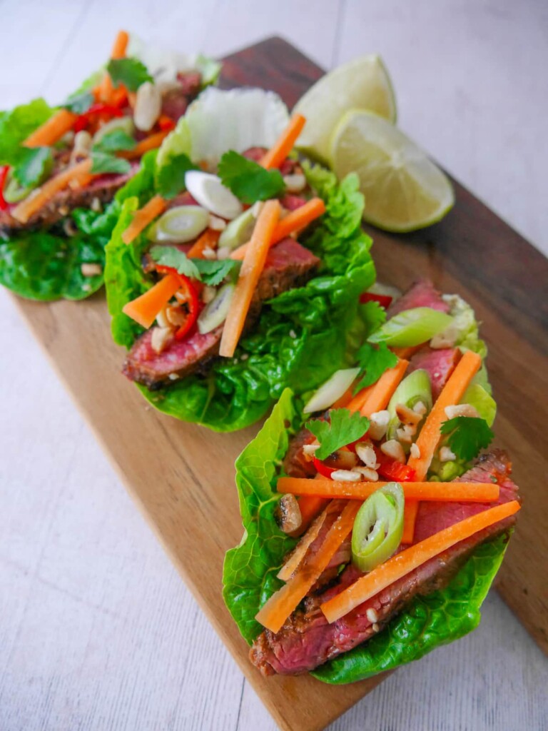 A wooden board topped with 3 lettuce leaves, laden with sliced lemongrass steak strips, crunchy carrot strips, spring onion, crushed peanuts and coriander leaf with wedges of fresh lime.