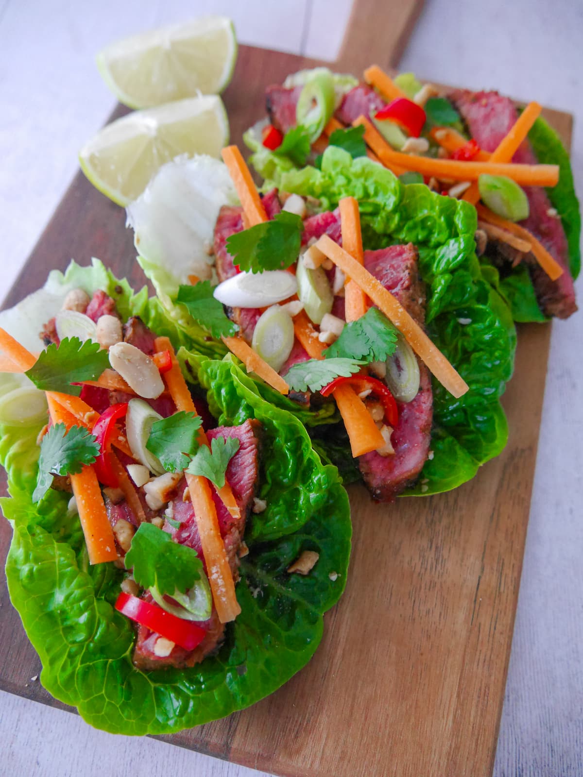 A wooden board topped with 3 lettuce leaves, laden with sliced lemongrass steak strips, crunchy carrot strips, spring onion, crushed peanuts and coriander leaf with wedges of fresh lime.