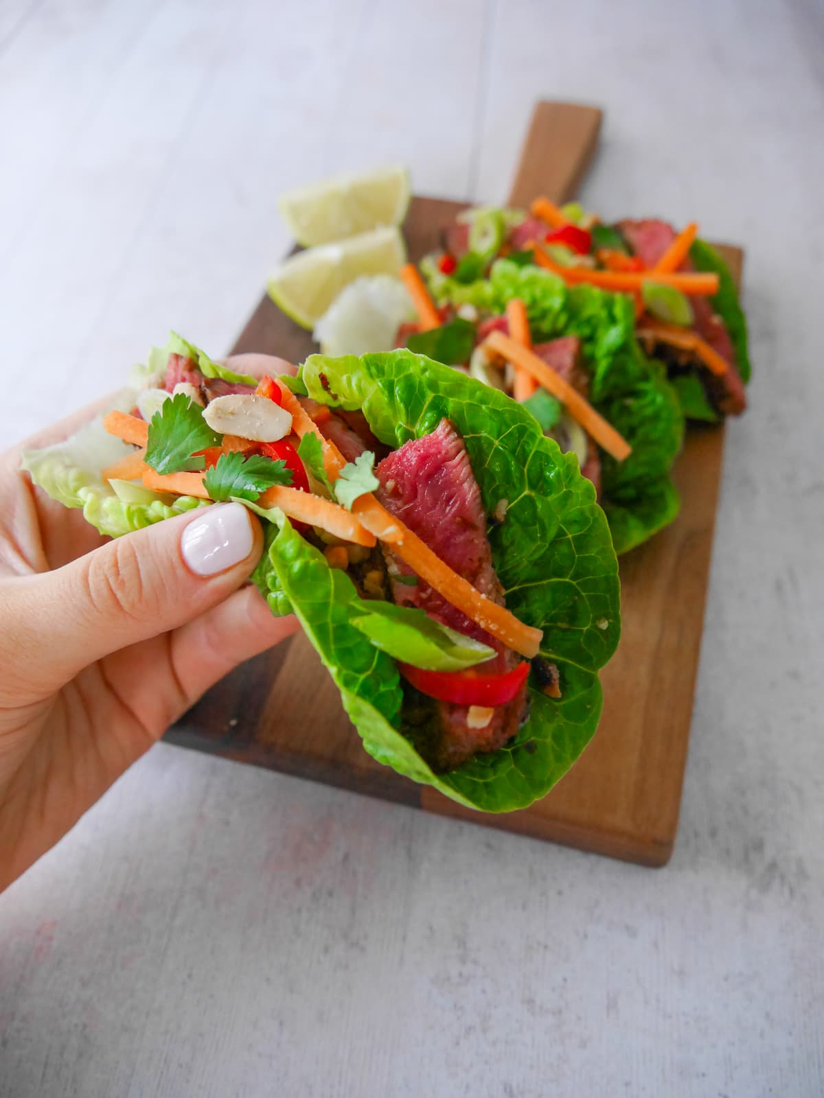 A hand holding a lettuce wrap laden with sliced lemongrass steak strips, crunchy carrot strips, spring onion, crushed peanuts and coriander leaf with wedges of fresh lime.