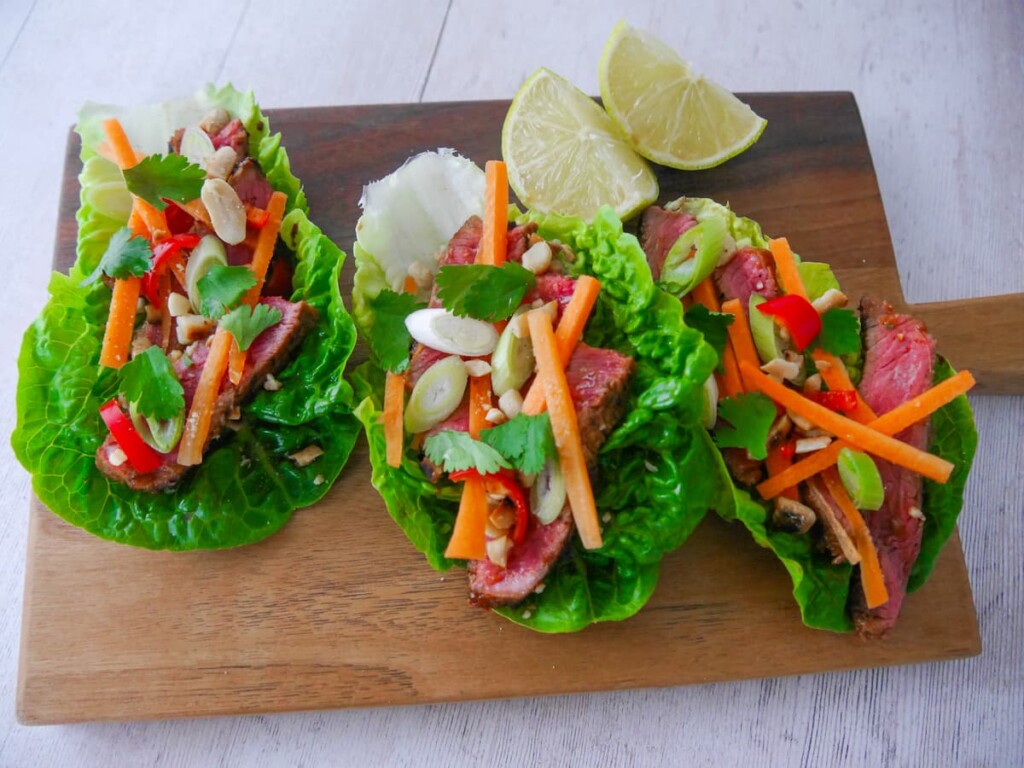 A wooden board topped with 3 lettuce leaves, laden with sliced lemongrass steak strips, crunchy carrot strips, spring onion, crushed peanuts and coriander leaf with wedges of fresh lime.